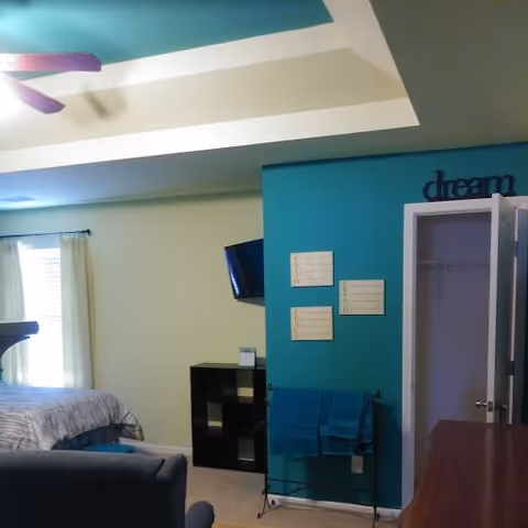 Interior view of a senior living facility bedroom with a bed, a ceiling fan, a small black shelving unit, a wall-mounted TV, an open closet door, and a teal accent wall with decorative signs and towels hanging on a rack.