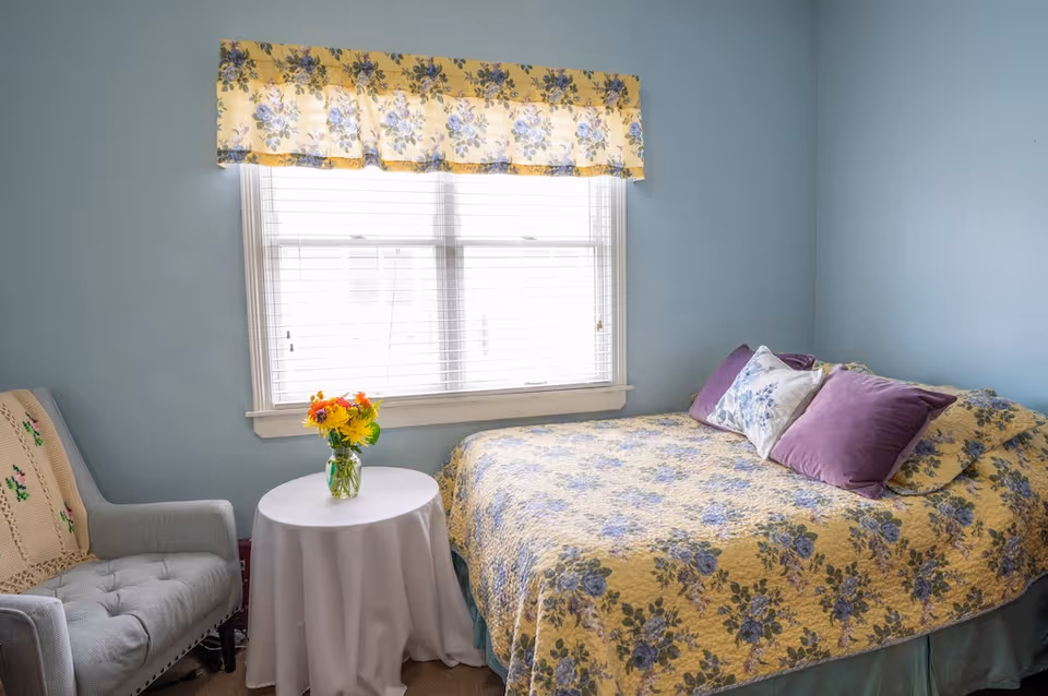 A cozy bedroom with light blue walls featuring a bed covered with a yellow floral quilt and purple and white pillows. Next to the bed is a small round table with a white tablecloth and a vase of colorful flowers. A gray upholstered armchair with a crocheted blanket is positioned beside the table. A window with white blinds and a yellow floral valance lets in natural light.