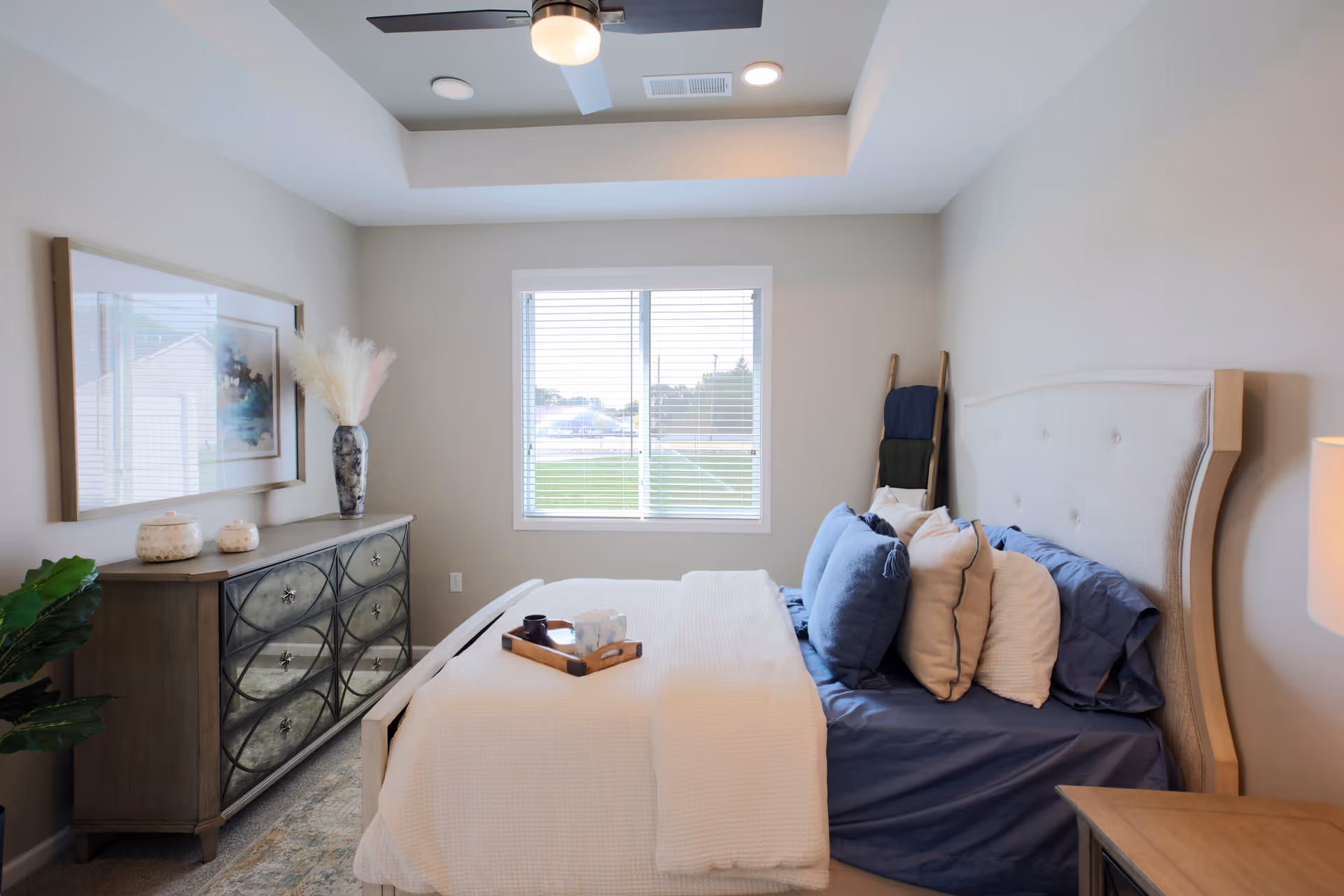 A neatly made bed with multiple pillows in shades of blue and white in a bright bedroom. The room features a window with blinds, a ceiling fan with light, a decorative dresser with a vase and artwork above it, and a small wooden tray on the bed. The walls are painted light gray, and there is a small ladder with blankets in the corner.