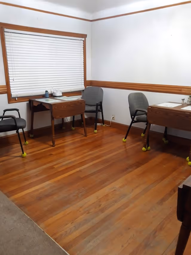 A small room with wooden floors and white walls featuring wooden trim. There are three chairs with tennis balls on the legs and two small wooden tables with placemats and a few items on top. A window with closed white blinds is on one wall.