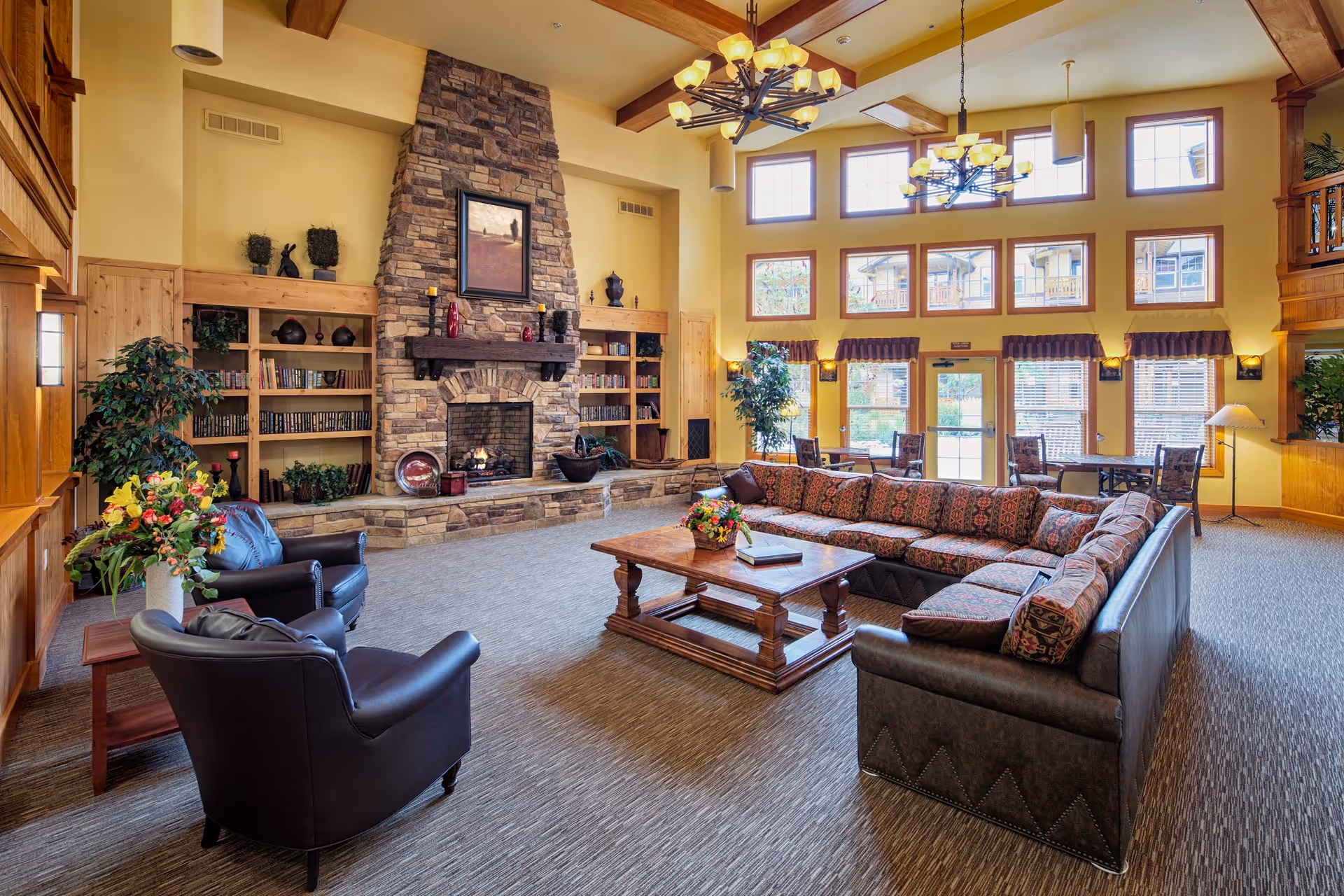 A spacious and well-lit living room area in a senior living facility featuring a large stone fireplace with a painting above it, built-in wooden bookshelves on either side, a large sectional sofa with patterned cushions, two black leather armchairs, a wooden coffee table with a flower arrangement, and tall windows letting in natural light. The room has warm yellow walls, wooden beams on the ceiling, and multiple chandeliers.