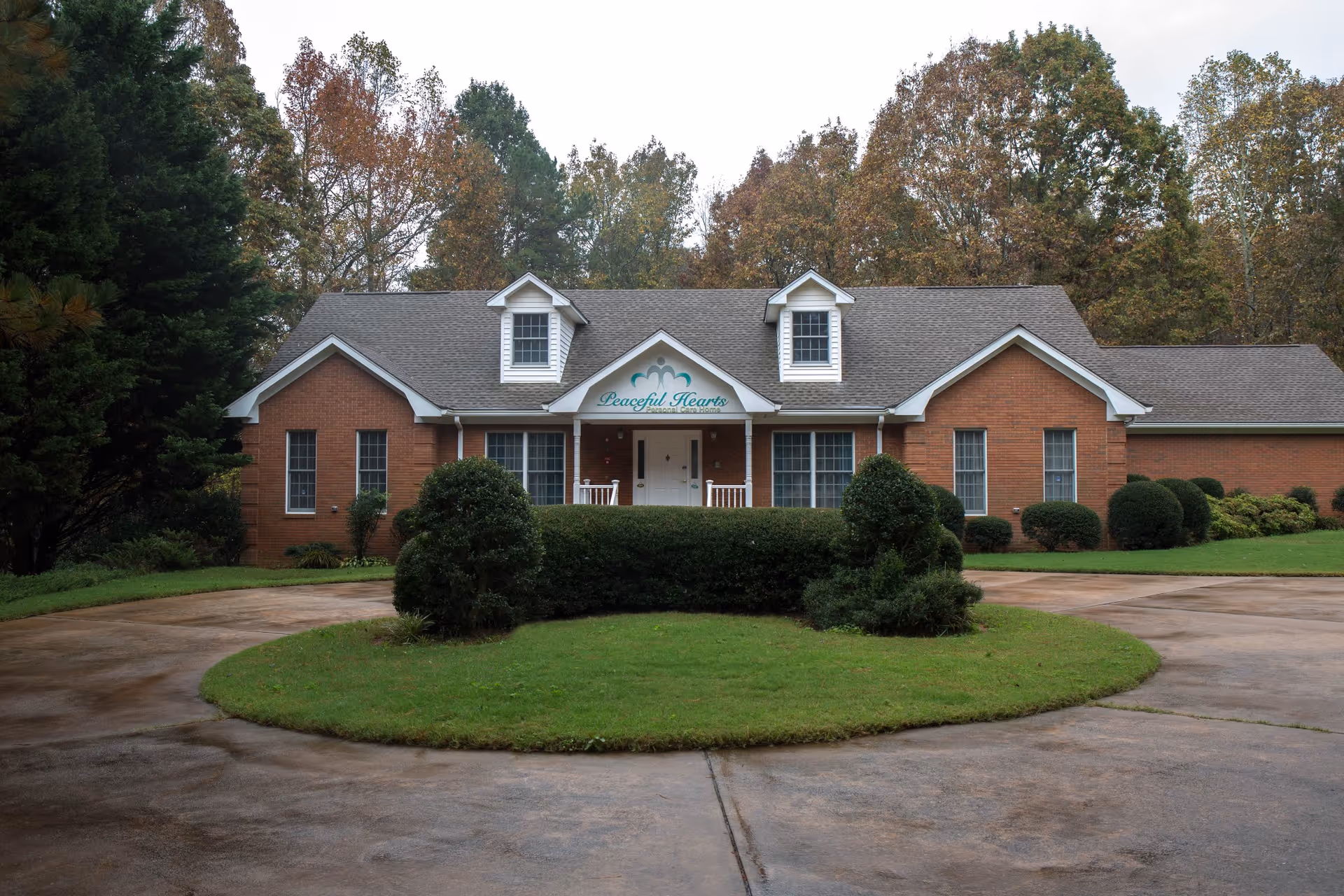 Front exterior view of a single-story brick building with a gray shingled roof, white trim, and two dormer windows. The building has a covered porch with white railings and a sign above the entrance that reads 'Peaceful Hearts Personal Care Home.' The driveway forms a circular loop around a grassy area with neatly trimmed bushes and trees in the background.