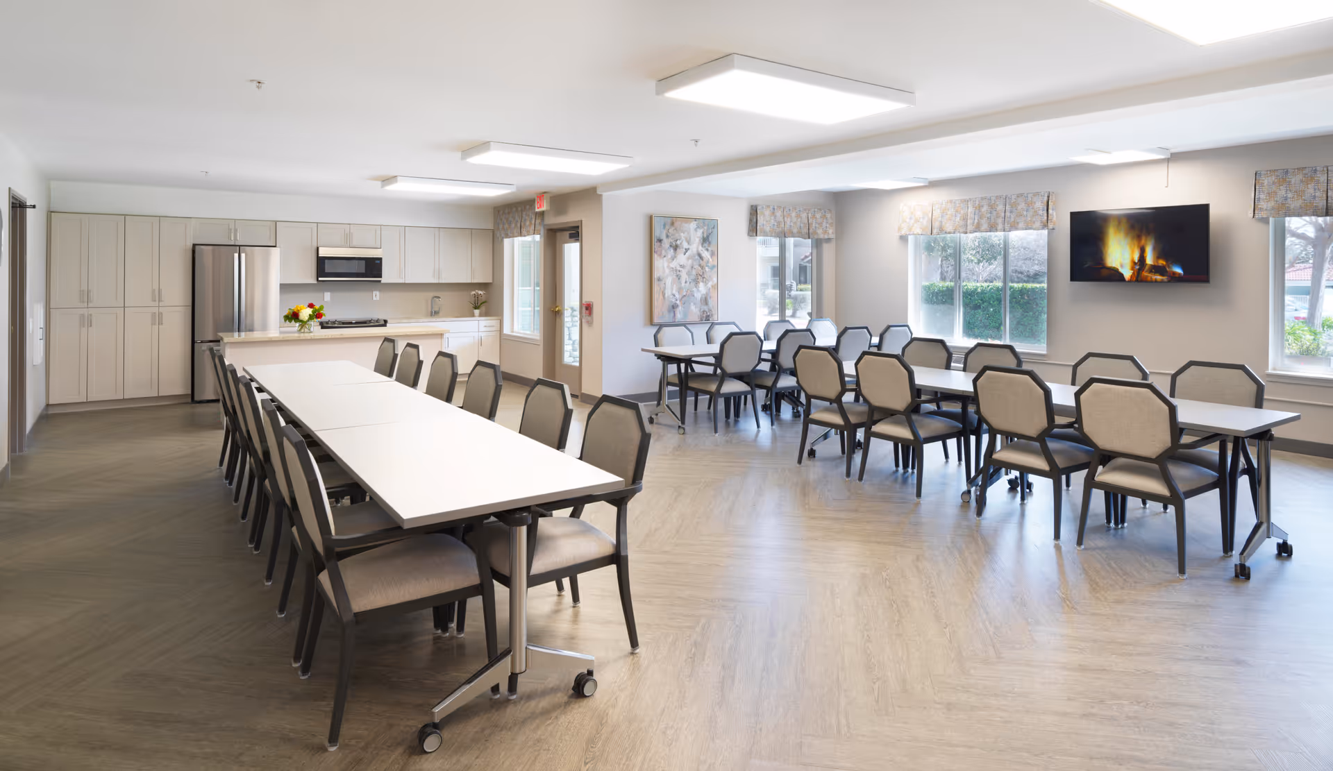 A bright and spacious communal dining area with two long tables surrounded by chairs. The room features a modern kitchen with white cabinets, a stainless steel refrigerator, and a microwave. Large windows with patterned valances allow natural light to fill the space. A flat-screen TV mounted on the wall displays a fireplace scene, and a colorful abstract painting hangs on one wall.