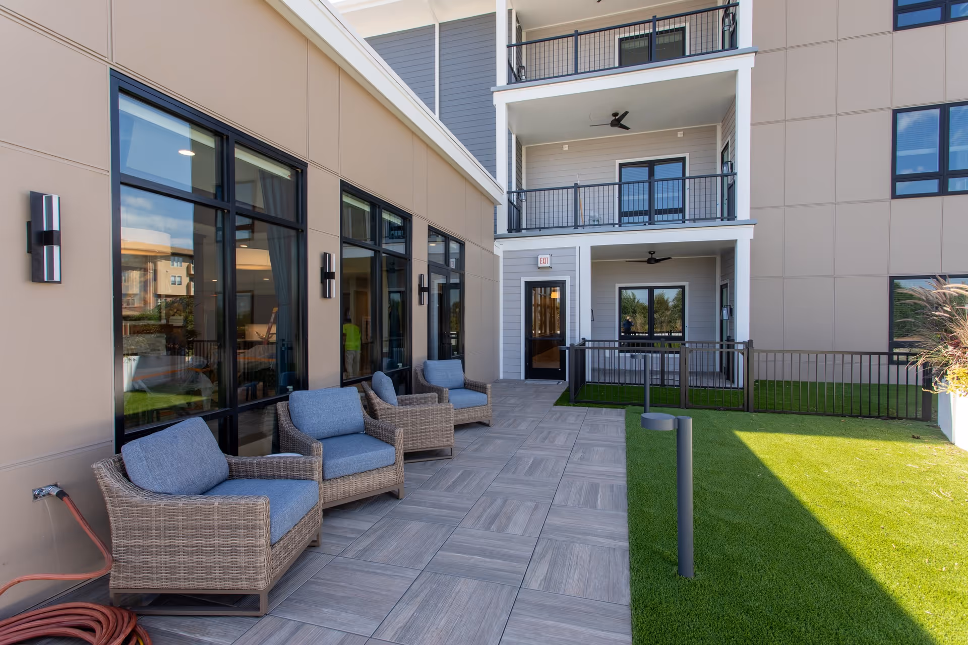 Outdoor patio area at Meadowlark Hills Retirement Community with four wicker chairs with blue cushions arranged along a building wall with large windows. The patio has tiled flooring and is adjacent to a green lawn area. The building has balconies with black railings and ceiling fans.