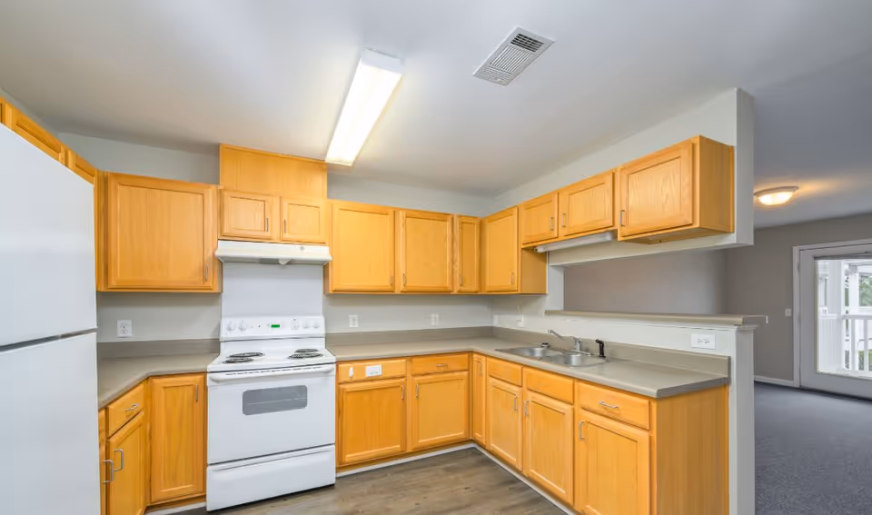 A bright kitchen with light wood cabinets, a white electric stove and oven, a white refrigerator, and a double sink. The countertops are a neutral gray color, and the floor is wood. There is a fluorescent ceiling light and a vent on the ceiling. The kitchen opens to a living area with carpet and a glass door leading outside.