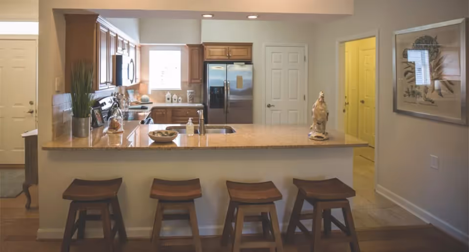 Open kitchen with a granite island and four wooden stools, stainless steel appliances, and oak cabinets.