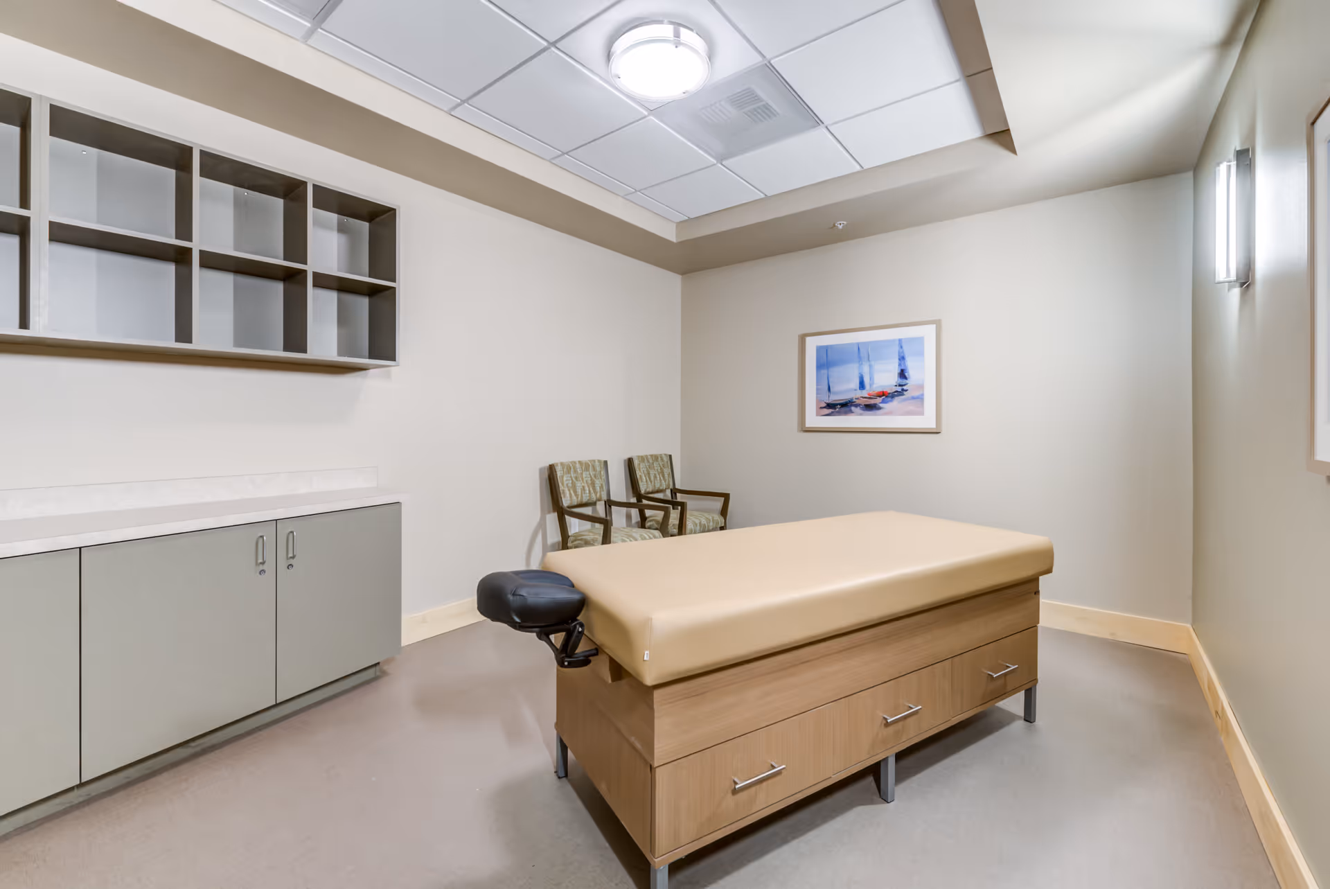 A small medical examination room with a beige padded examination table in the center, two patterned chairs against the wall, a cabinet with drawers and open shelving, and a framed picture of sailboats on the wall.