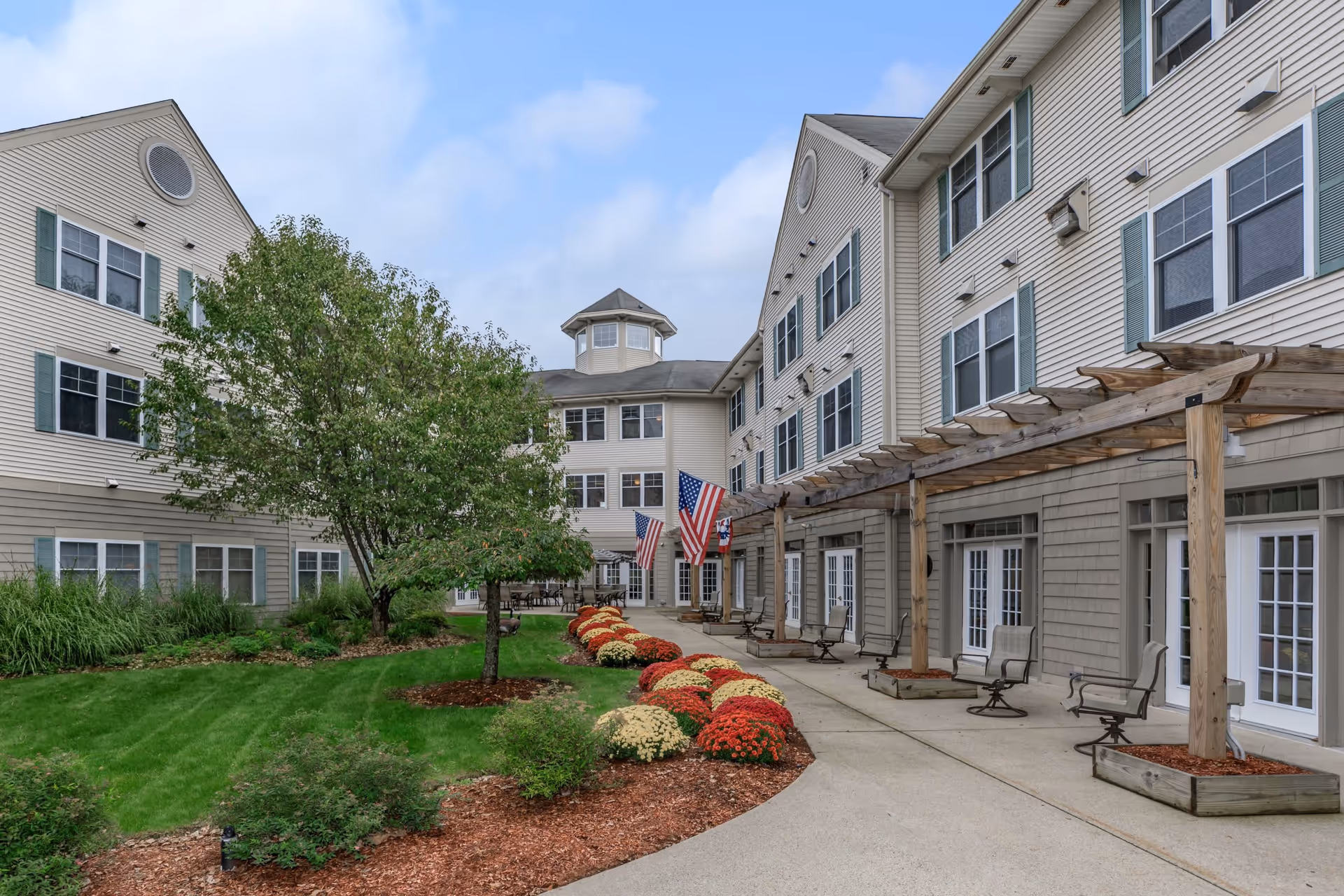 Outdoor courtyard area of a senior living facility with a well-maintained lawn, flower beds with red and white flowers, a tree, and a paved walkway. The building has multiple windows with green shutters, a wooden pergola with chairs underneath, and American flags displayed near the entrance.