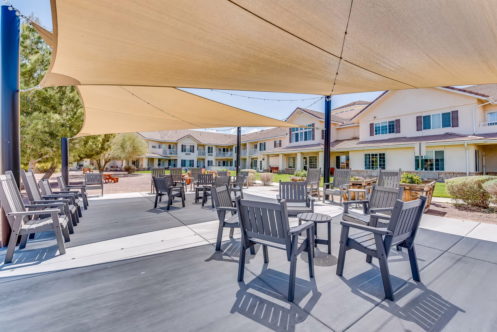 Outdoor seating area at a senior living facility with multiple gray chairs and small tables arranged under large beige shade sails. The background shows a two-story building with windows and balconies, surrounded by trees and shrubs under a clear blue sky.