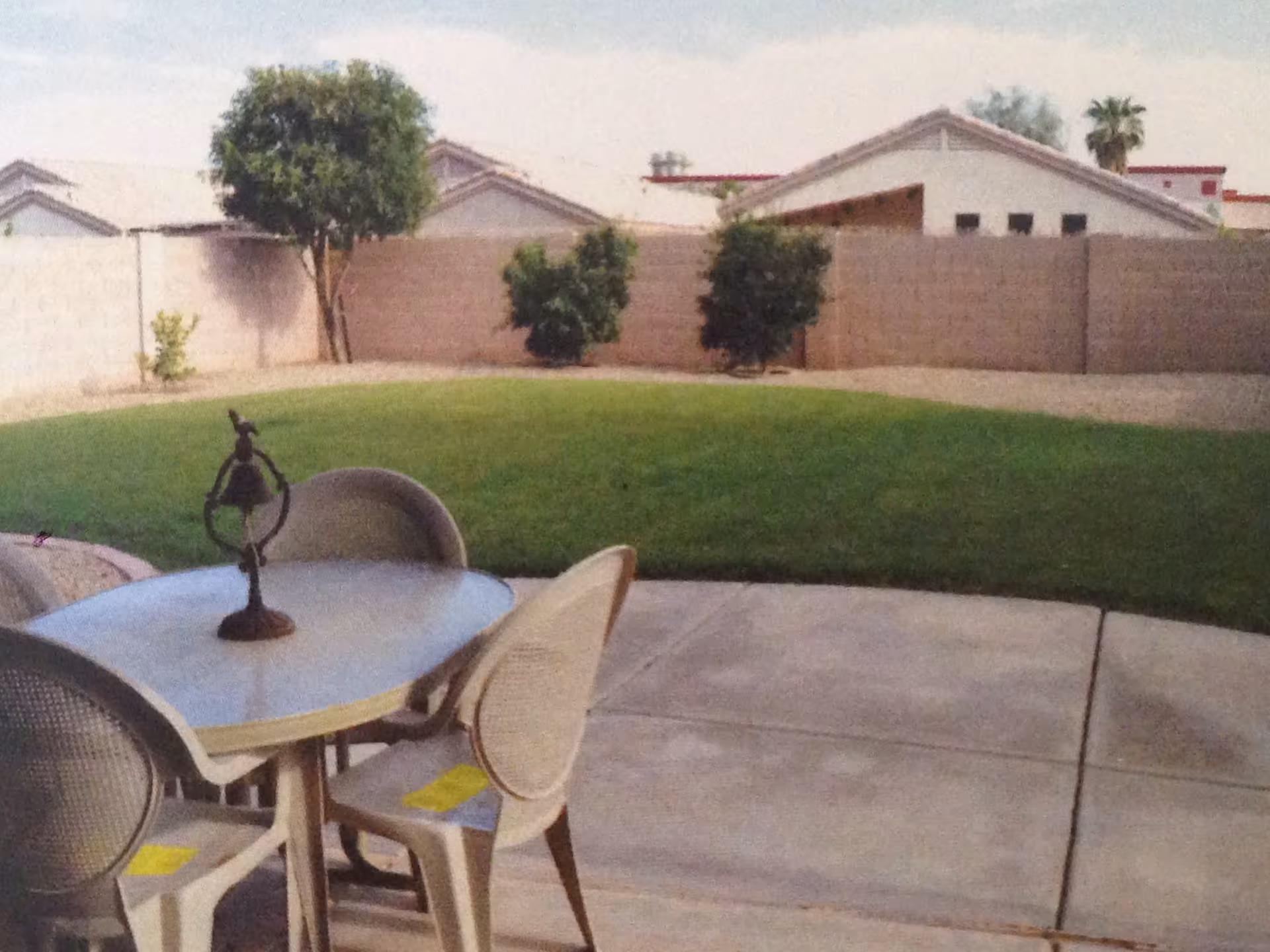 Outdoor patio area with a round table and four chairs on a concrete surface, overlooking a green lawn with a few small trees and bushes, surrounded by a beige brick wall and neighboring houses in the background.