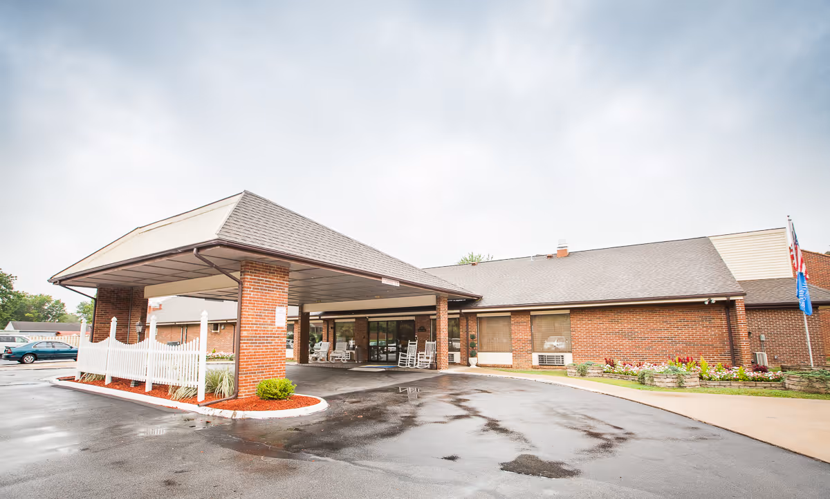 Exterior view of a single-story brick building with a covered entrance and a driveway in front. There are rocking chairs under the covered entrance, a small landscaped area with flowers and shrubs, and flags on poles to the right side of the building. The sky is cloudy.