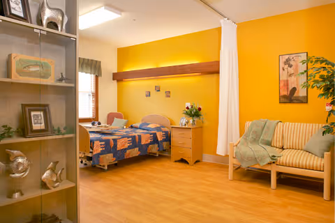 A cozy bedroom in a senior living facility with a single bed covered in a colorful quilt, a wooden nightstand with flowers, a small couch with a green throw blanket, a tall glass display cabinet with decorative items, and a bright yellow accent wall.