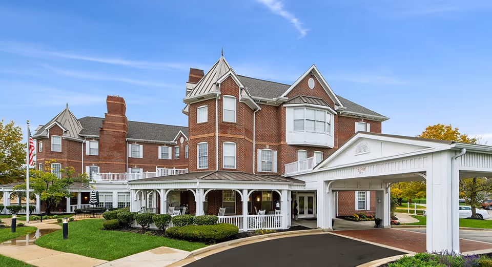 Exterior view of a large, multi-story brick building with white trim and a covered entrance. The building has multiple windows, a turret-like structure, and a well-maintained lawn with shrubs and a flagpole displaying the American flag. The sky is clear and blue.
