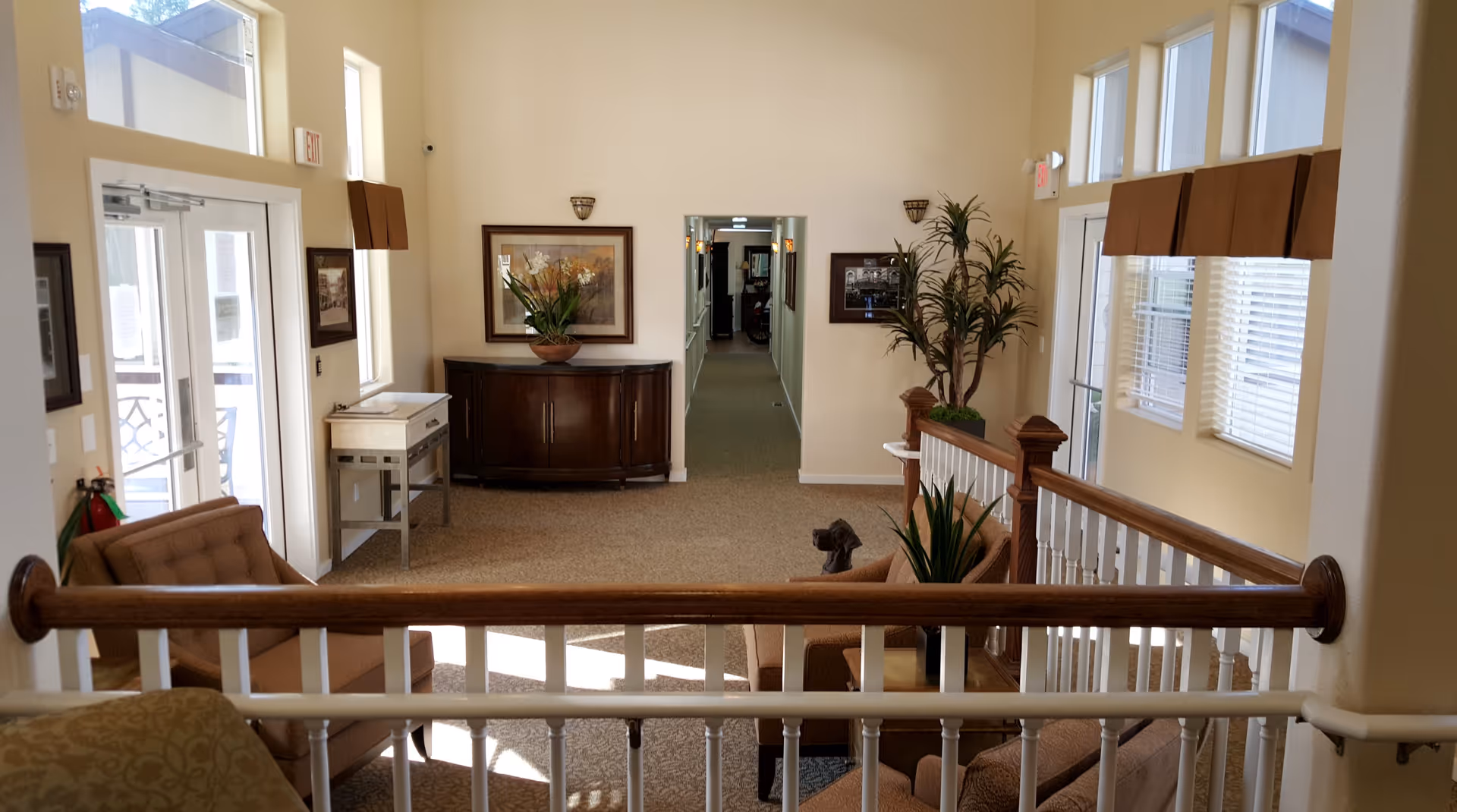 Interior view of a well-lit common area in a senior living facility with beige walls and carpet. The space features comfortable brown upholstered chairs, a wooden railing, a dark wooden sideboard with a flower arrangement, framed artwork on the walls, and large windows with brown valances. A hallway extends from the center of the room, and there are exit signs above the doors.
