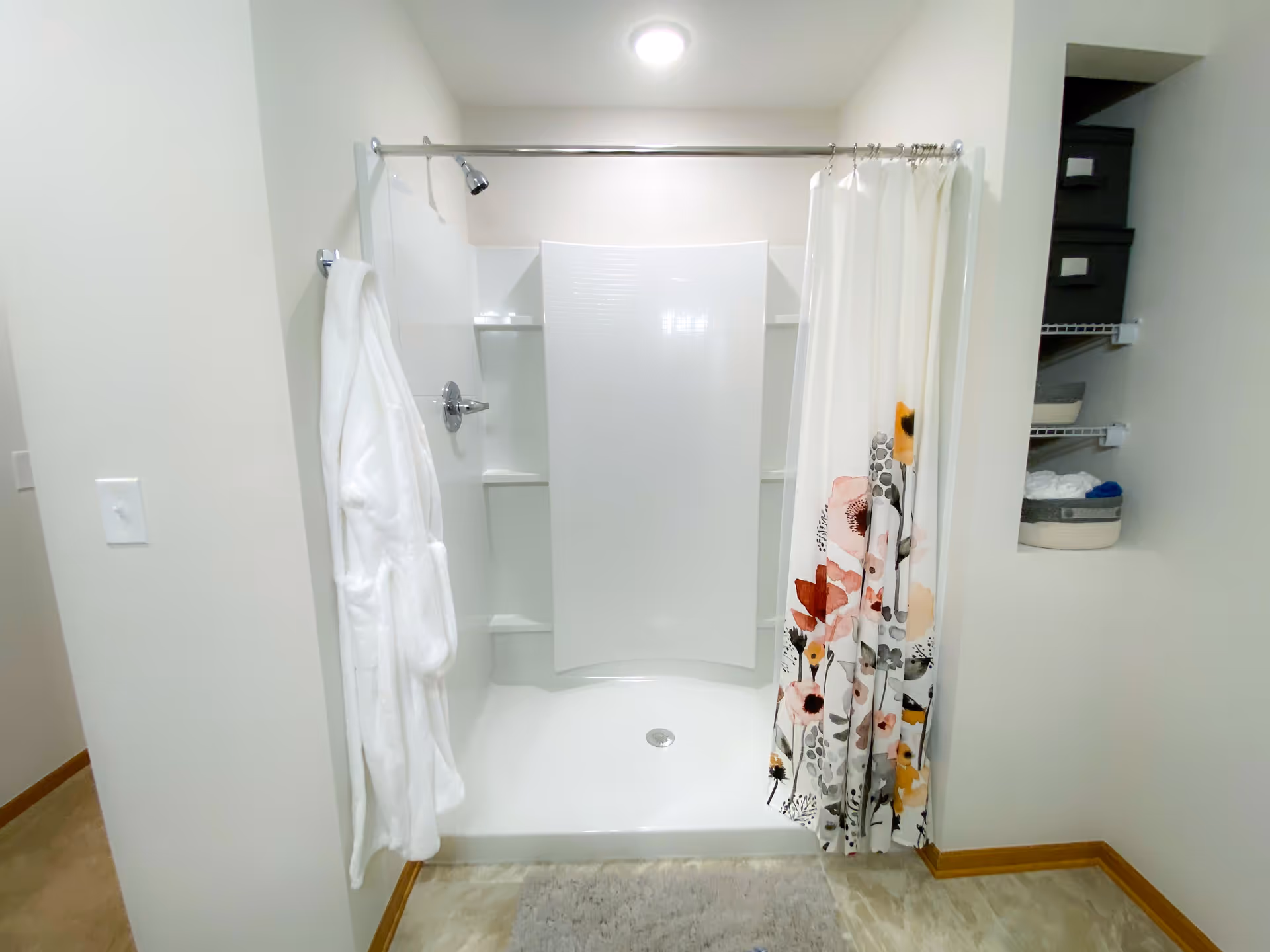 A clean, white shower stall with a floral-patterned shower curtain partially drawn to the right. A white bathrobe hangs on the left wall next to the shower. To the right, there are open shelves with storage baskets and folded towels. The floor has a light-colored mat in front of the shower.
