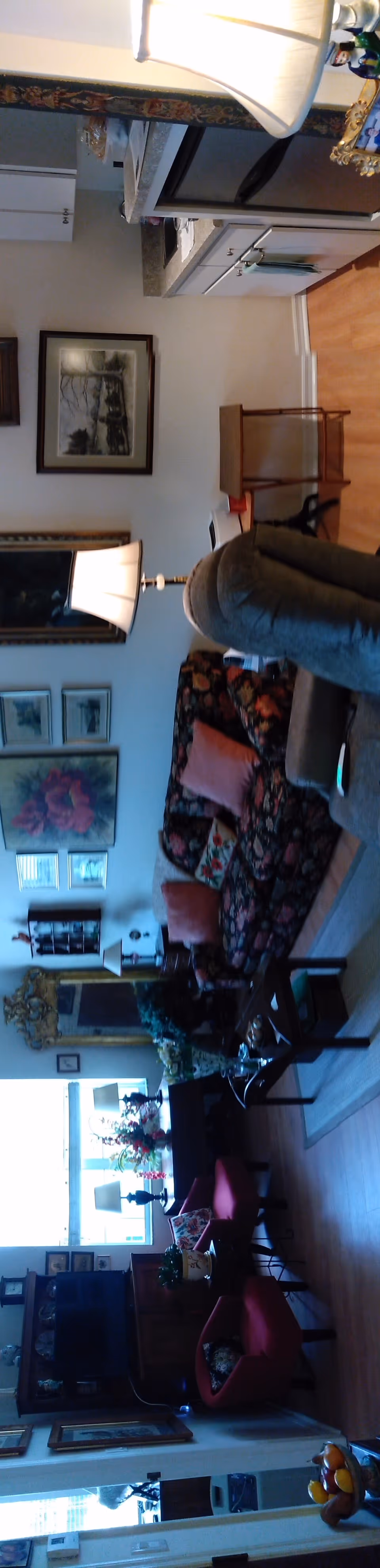 A cozy living room area with floral patterned sofa and multiple cushions, a wooden coffee table, two red chairs, a floor lamp, framed artwork on the walls, and a small kitchen area with a countertop and cabinets in the background.