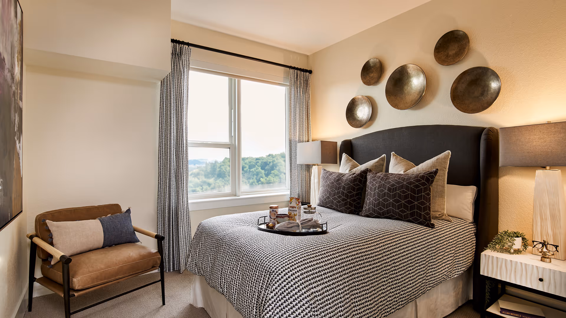 A cozy bedroom with a neatly made bed, patterned bedding and pillows, metal wall plates above the headboard, bedside lamps, an armchair, and a window overlooking trees.