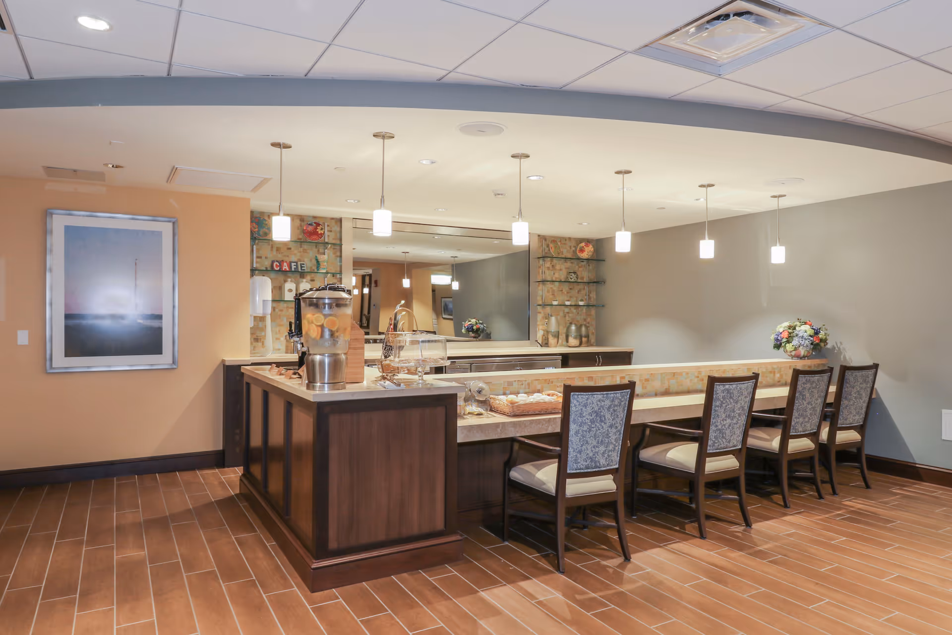 A modern cafe-style seating area with a long counter and five chairs with patterned upholstery. The counter has a beverage dispenser with lemon water, a basket of pastries, and decorative items. The background features a large mirror, shelves with decorative plates, and pendant lights hanging from the ceiling. The floor is tiled with wood-like tiles, and the walls are painted in neutral tones with a framed picture on one side.