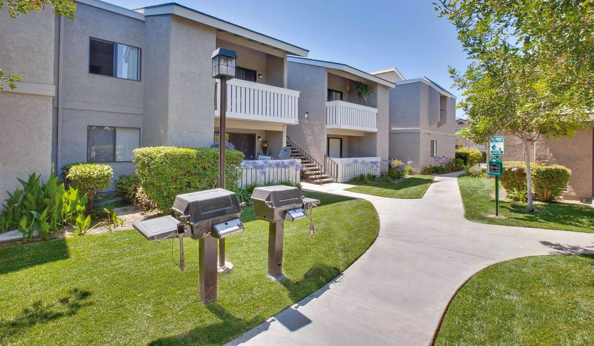 Two outdoor barbecue grills on a grassy courtyard in front of a two-story apartment building with balconies and a paved walkway.