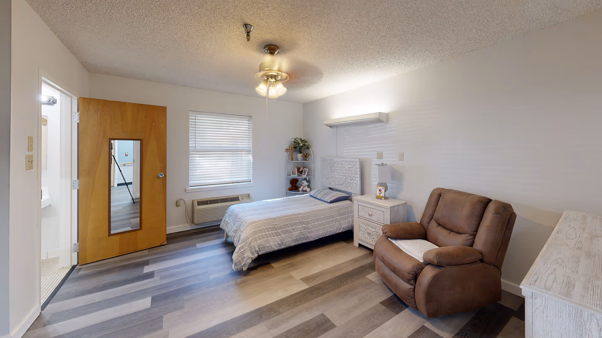A simple senior living bedroom with a single bed, nightstand, recliner chair, and a window with blinds.