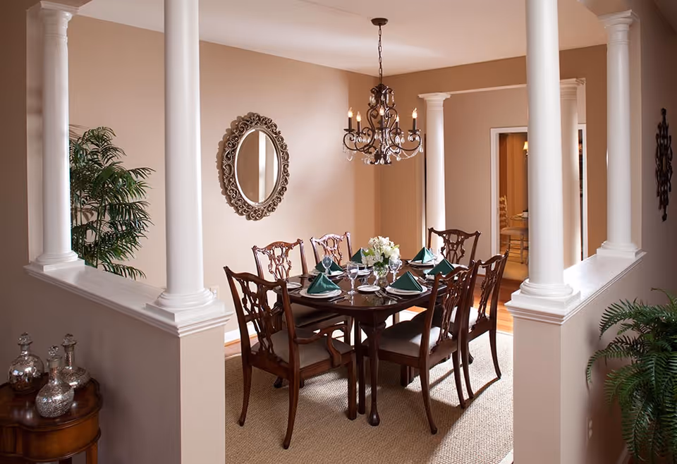 A formal dining room with a dark wooden dining table set for six people with green folded napkins, white plates, and glassware. The room features beige walls, white decorative columns, a chandelier hanging above the table, a round ornate mirror on the wall, and green plants in the corners.