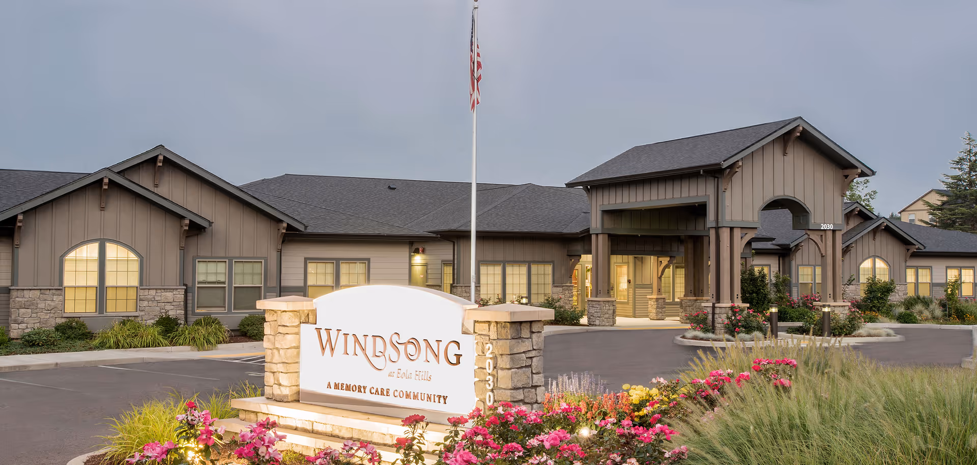 Front exterior of the Windsong at Eola Hills memory care community showing the building entrance, sign, flagpole, and landscaped flowers.