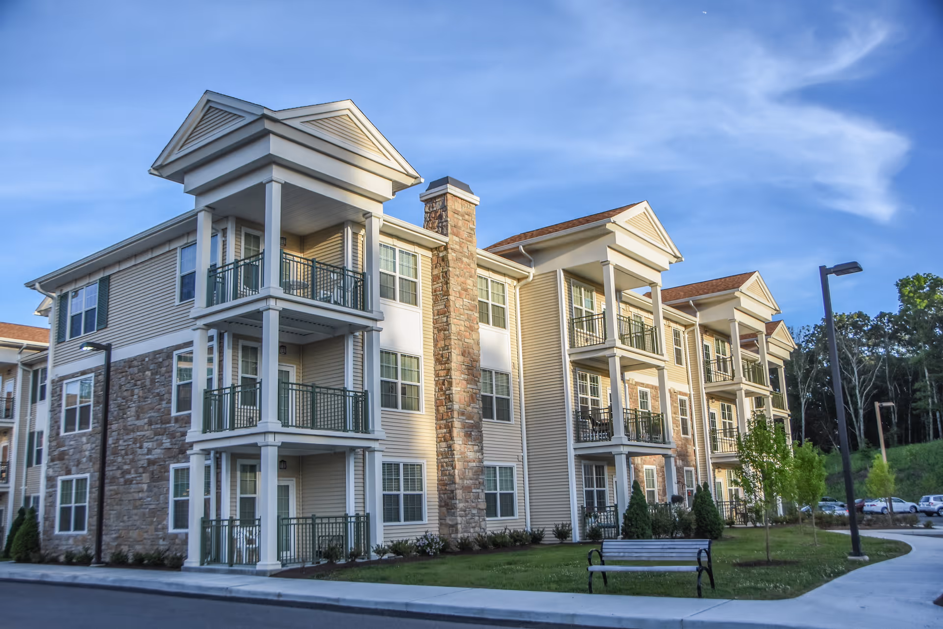 Front exterior of a three-story beige apartment-style senior living building with balconies, stone accents, a bench, and lamp posts under a blue sky.