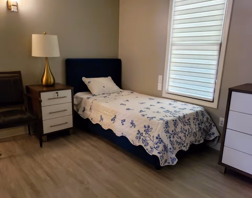 A small bedroom with a single bed covered with a white bedspread featuring blue floral patterns. Next to the bed is a wooden nightstand with three drawers and a gold-colored lamp with a white lampshade. There is a dark chair beside the nightstand. A window with a white blind is on the right wall, and a white dresser with dark edges is partially visible on the right side of the image. The floor is light wood, and the walls are painted beige.