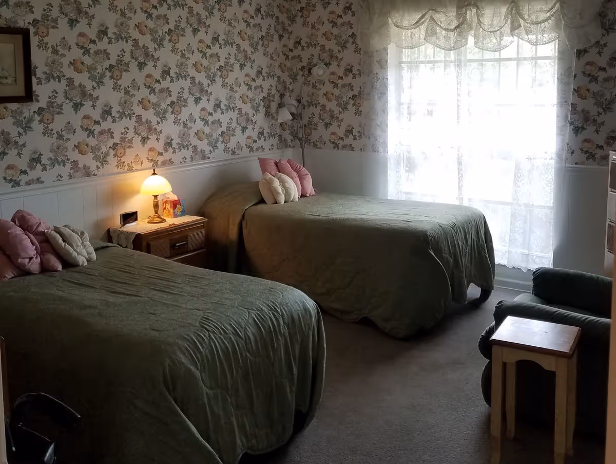 A bedroom with two twin beds covered in green bedspreads, a bedside table with a lamp, floral wallpaper, and a lace-curtained window.