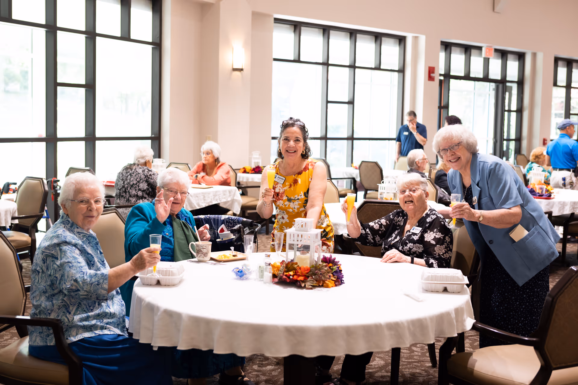 A group of elderly women and a staff member sitting and standing around a round table in a bright dining room, smiling and holding drinks. The table is decorated with a centerpiece and has food containers and cups on it. Other elderly people are visible in the background seated at tables near large windows.