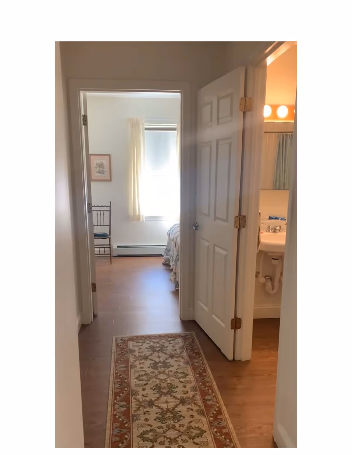 View down a hallway with a patterned runner rug on a wooden floor. On the right, an open door reveals a bathroom with a sink and mirror. Straight ahead, an open doorway leads to a bedroom with a window, light curtains, a chair, and part of a bed visible.