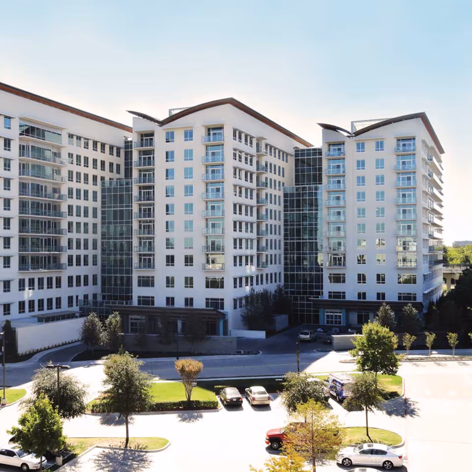 Exterior front view of a multi-story white senior living building with balconies, a central glass atrium, and a parking area with trees.