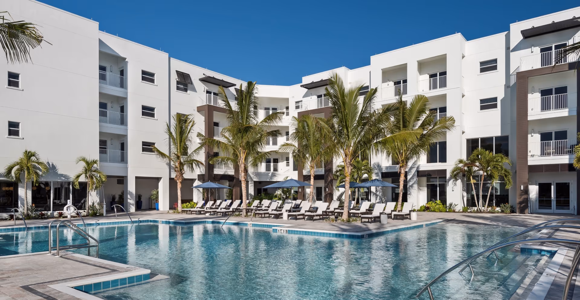 Outdoor swimming pool with lounge chairs and palm trees in the courtyard of a white multi-story senior living building.