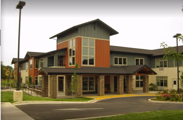 Front exterior of a two-story assisted living facility with a covered entrance supported by stone pillars, large windows, and a landscaped driveway.