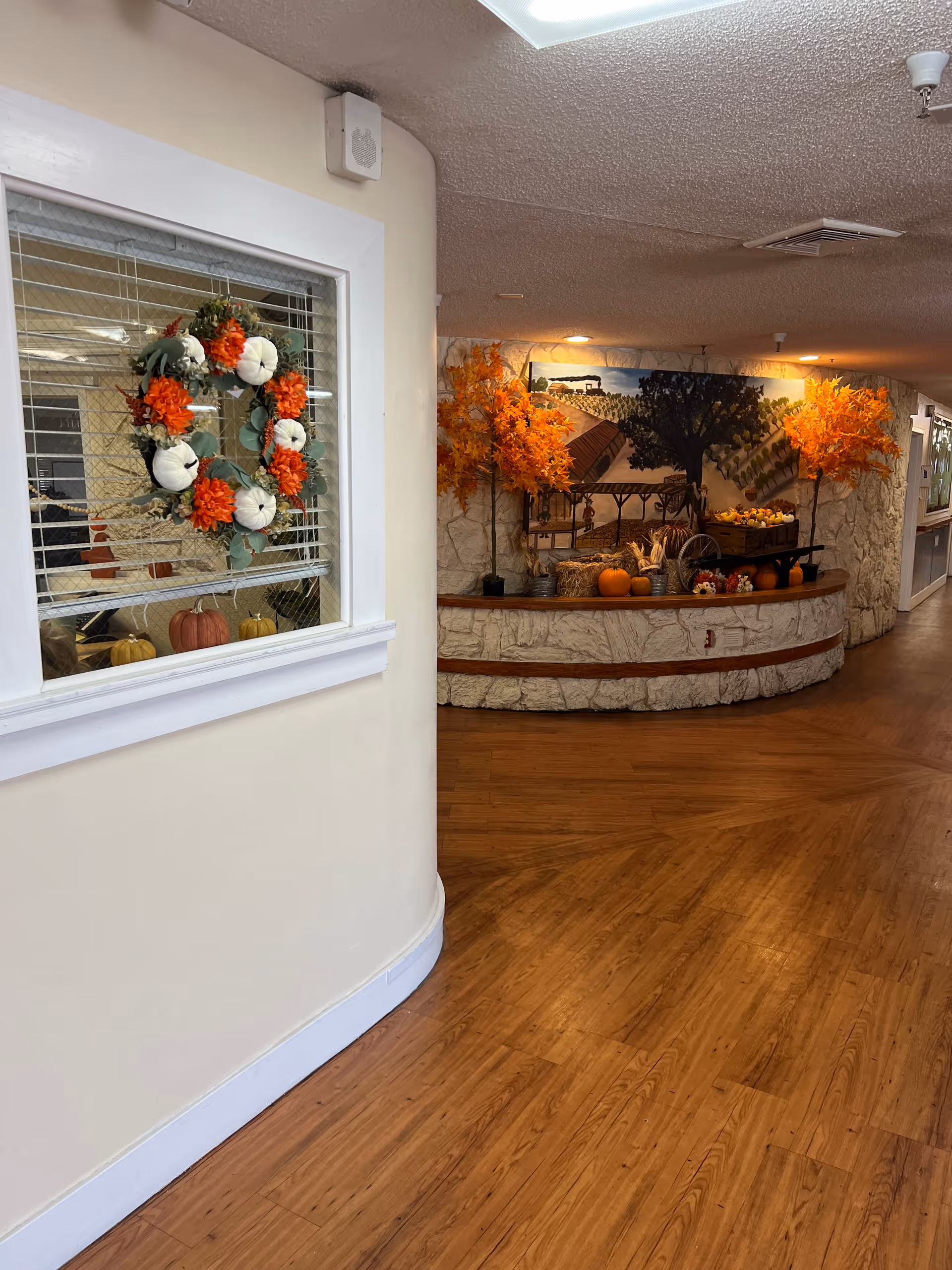 Interior hallway of a senior living facility decorated for autumn with orange and white pumpkin wreath on a window, two orange artificial trees, pumpkins, hay bales, and a mural depicting a vineyard scene on a stone-textured wall.