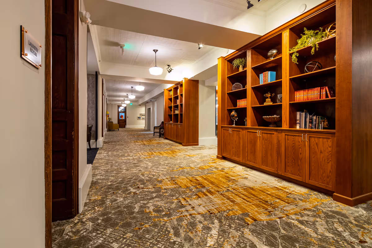 A well-lit hallway with patterned carpet and wooden bookshelves filled with books and decorative items. The hallway has ceiling lights and a sign on the left wall indicating a laundry room.