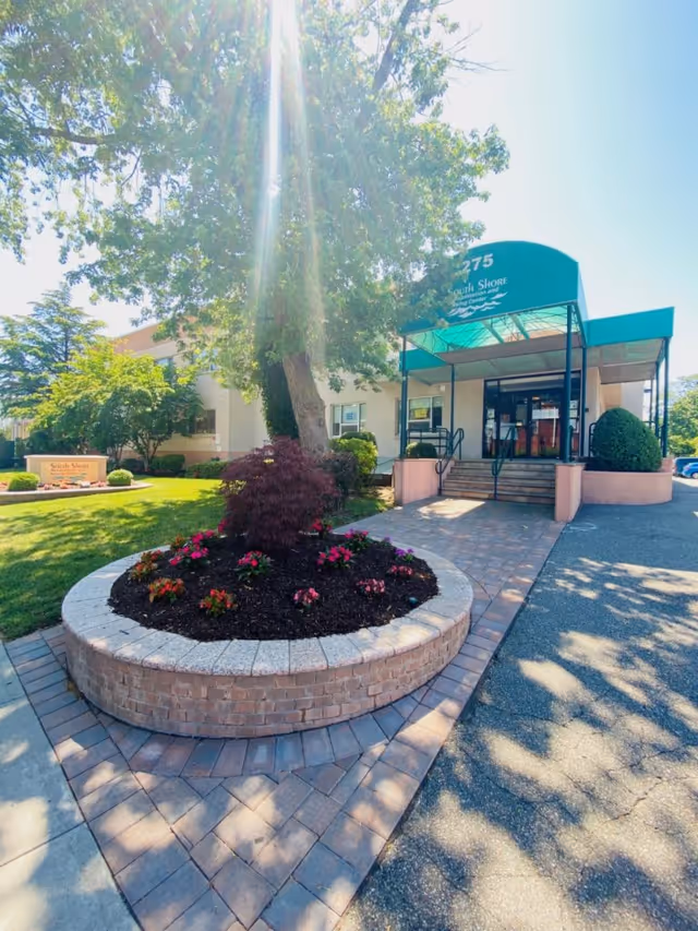 Front entrance of South Shore Rehabilitation and Nursing Center showing a covered entry with steps, a circular brick planter with flowers, and a large tree.