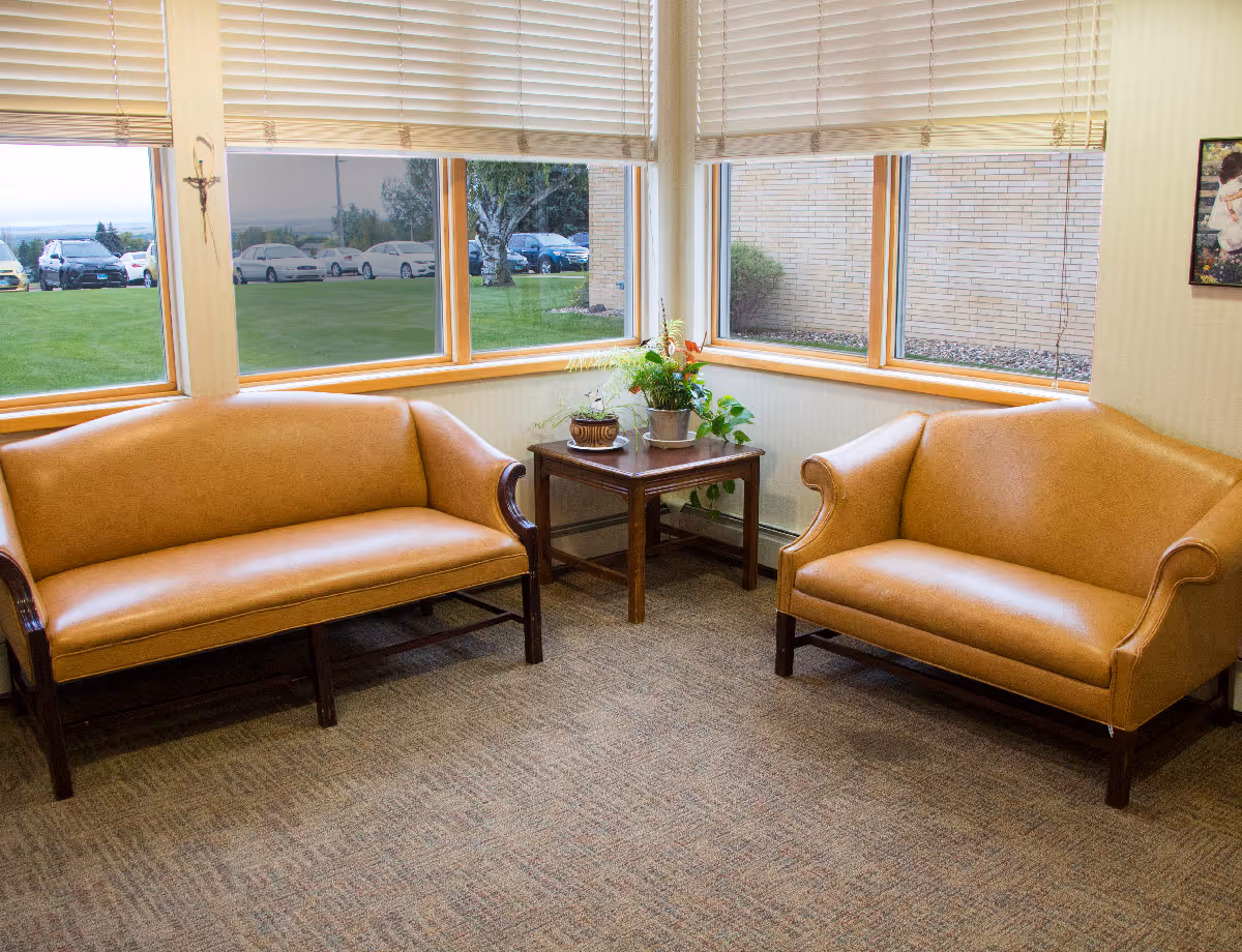 A cozy corner seating area with two tan leather sofas facing each other and a wooden side table with potted plants in between. Large windows with beige blinds provide a view of a green lawn, parked cars, and a brick building outside.