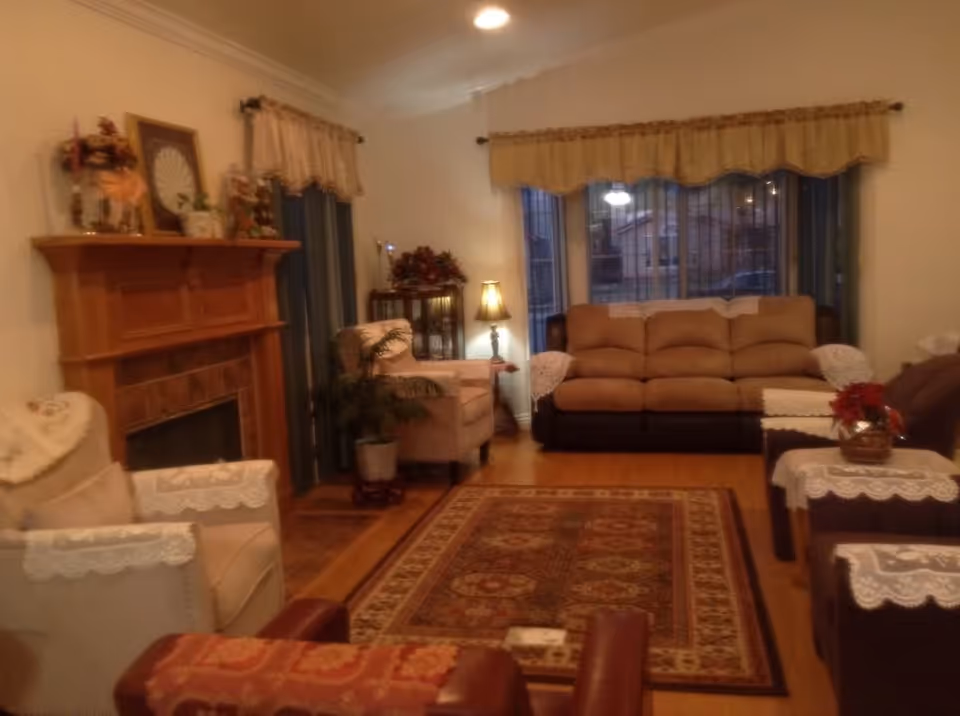 Cozy living room with a sofa and armchairs arranged around a patterned rug, a fireplace on the left, and a large window with valances.