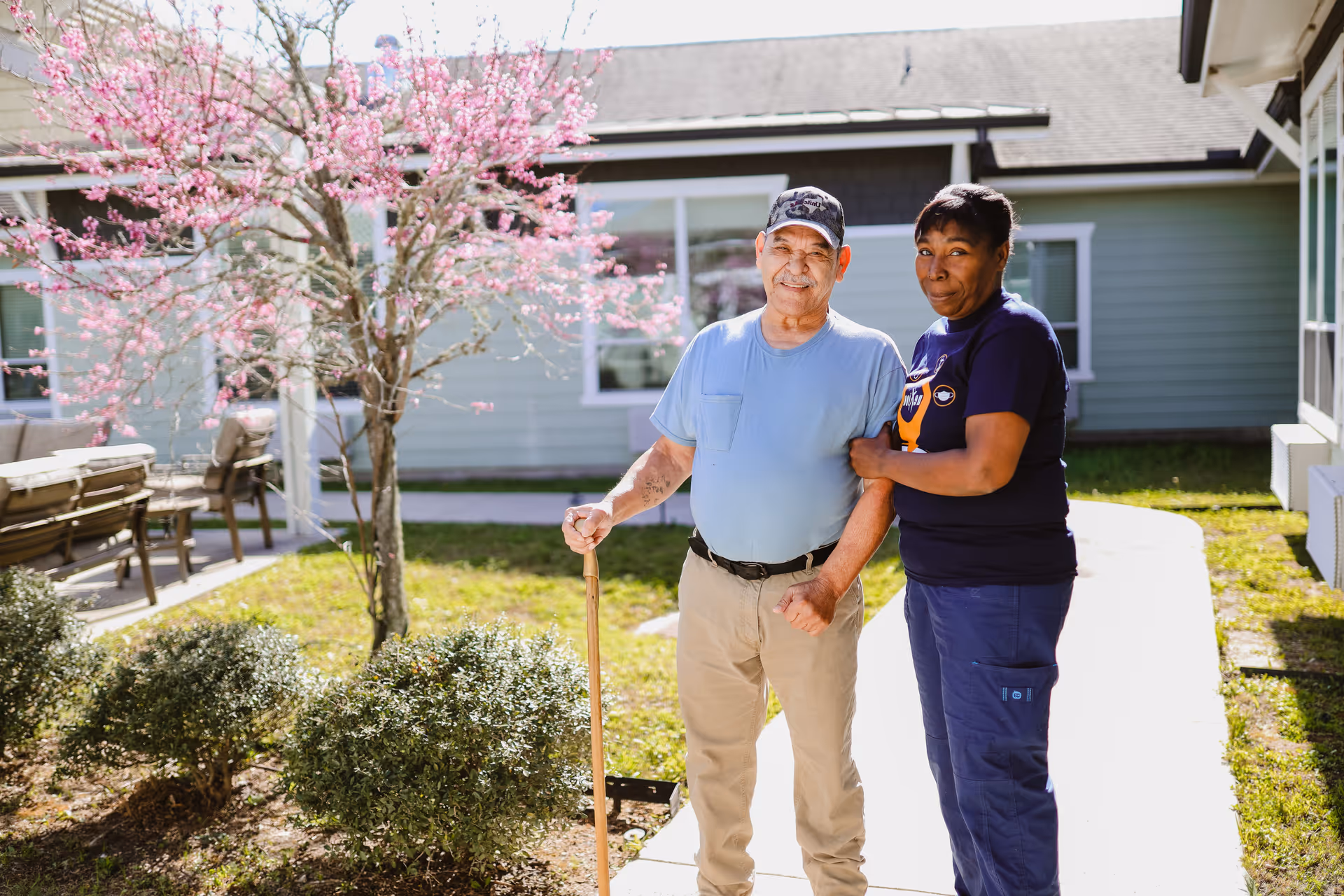 An elderly man with a walking cane stands outside on a sunny day next to a caregiver who is holding his arm. They are in a garden area with green bushes and a tree with pink blossoms, in front of a light green building with windows.