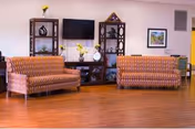 A living room area with two patterned orange sofas facing each other on a wooden floor. Between them is a dark wooden shelving unit with decorative items and a flat-screen TV mounted on the wall above it. There are framed pictures on the walls and a vase with yellow flowers on the shelving unit.