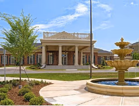 Front exterior view of The Woodlands at Furman facility featuring a large building with columns at the entrance, a circular driveway, landscaped greenery, and a multi-tiered water fountain in the foreground under a blue sky.
