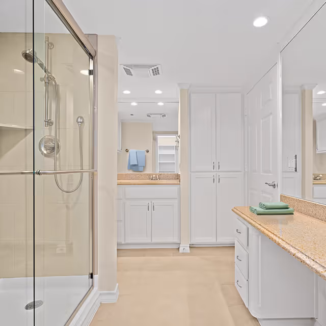 Bright and clean bathroom with a glass-enclosed shower on the left, white cabinetry, beige countertops, and a large mirror. A blue towel hangs on a rack near the sink, and the floor is tiled in a light beige color.