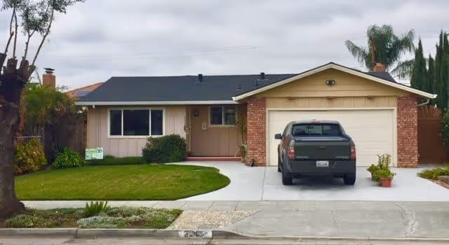Single-story residential house with a brick and beige exterior, a black roof, a driveway with a parked black pickup truck, a green lawn, and some plants and trees around the property under a cloudy sky.