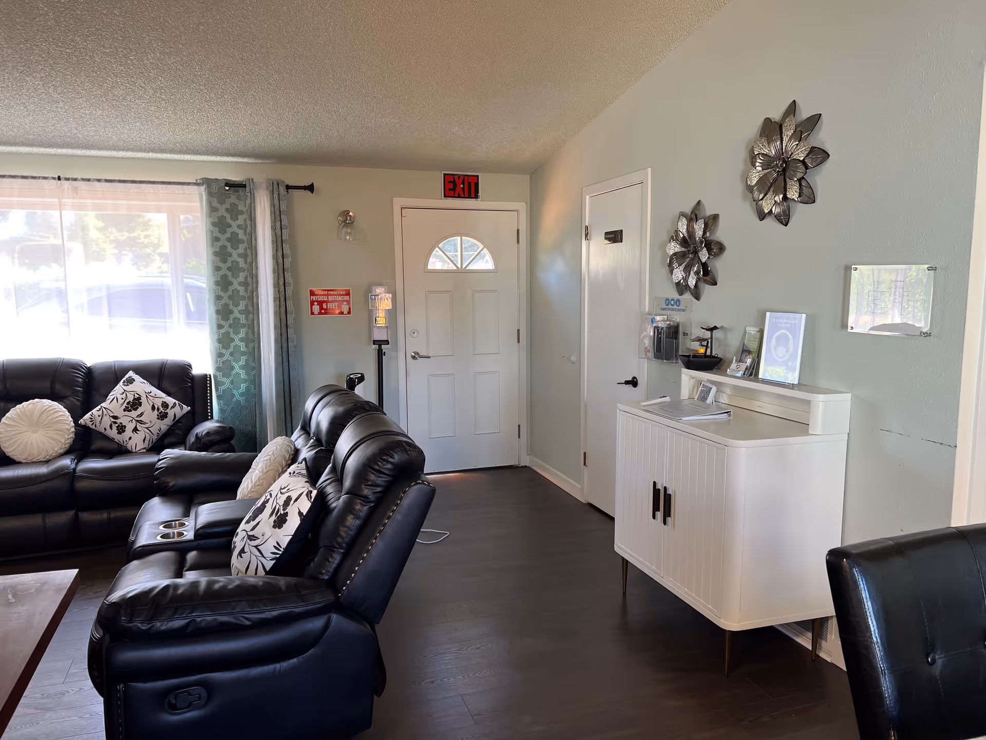 Interior view of a living room area with black leather recliner sofas adorned with patterned and round pillows. A large window with sheer and patterned curtains lets in natural light. A white door with a small window is visible in the background with an exit sign above it. On the right wall, there is a white cabinet with decorative items and two metallic flower wall decorations above it. The floor is dark wood.