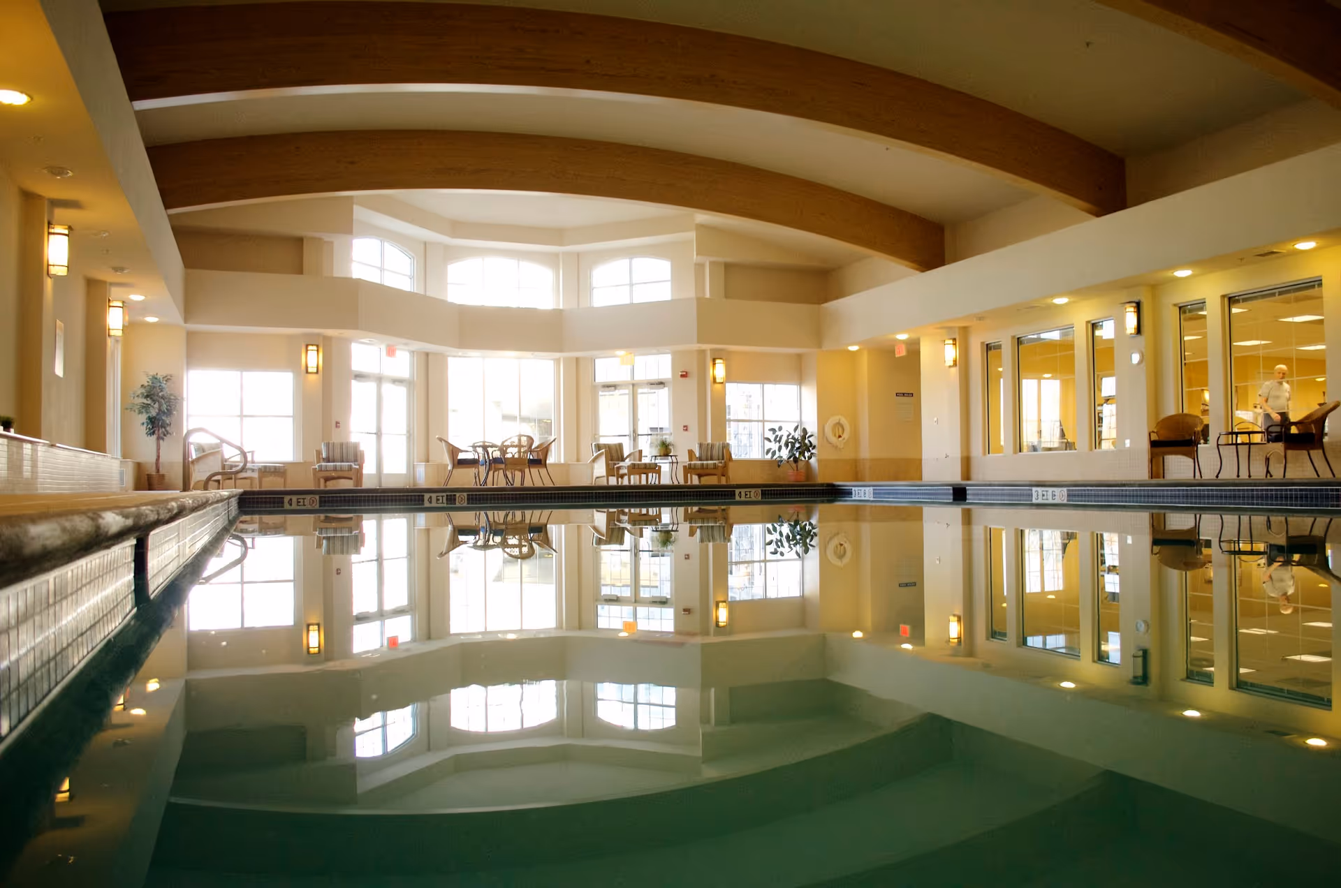 Indoor swimming pool area with calm water reflecting the ceiling and large windows. The room has wooden beams on the ceiling, several chairs and tables along the walls, and large windows letting in natural light. There is a person visible through the glass windows on the right side.