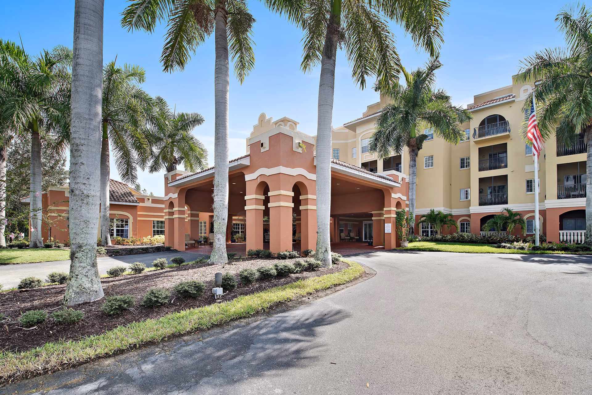 Front entrance of a Mediterranean-style senior living building with a covered porte-cochere, palm trees, and an American flag.