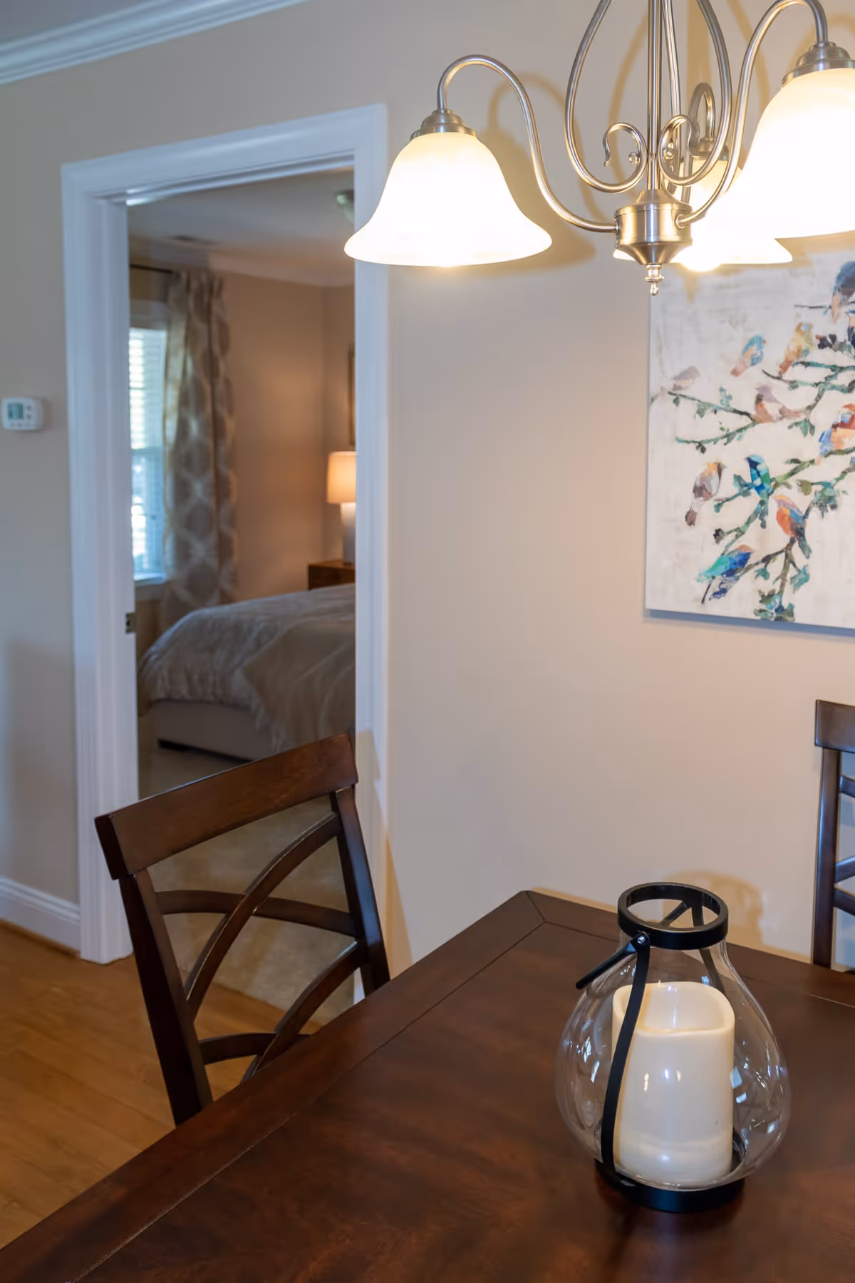 A dining area with a wooden table and chairs, a glass candle holder with a white candle on the table, a hanging light fixture with three lamps, and a wall painting of colorful birds on branches. In the background, there is a doorway leading to a bedroom with a bed, bedside lamp, and window with curtains.