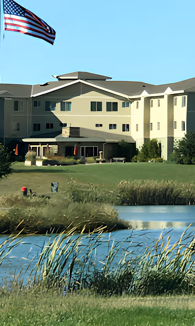 Multi-story senior living building with a lawn, pond in the foreground and an American flag flying above.