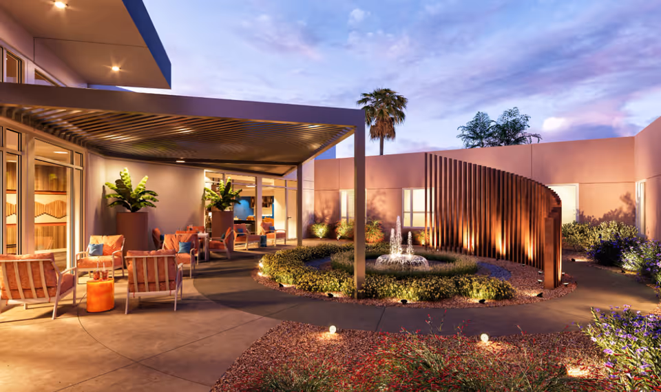 Outdoor courtyard area at dusk with seating arrangements under a pergola, decorative plants, a circular garden with a small water fountain, and modern architectural elements including a curved wooden screen and surrounding walls.